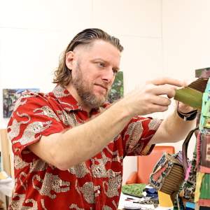 Augustus Hoffman, a white man with dark blonde hair and a beart, works on an abstract sculpture made from various materials.