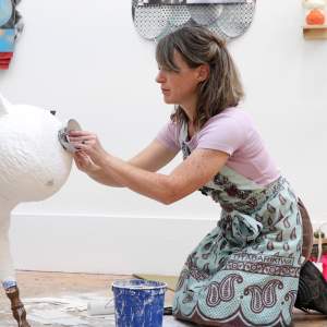 Emily Holt kneels in a studio as she applies papier mache to a small cow-like sculpture. She is a white woman with light skin tone and shoulder length dark blonde hair, wearing a patterned apron.