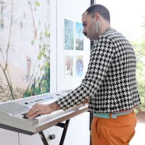 Lucia Honey plays an electronic keyboard in an art studio. She has light toned skin, a dark mustache, buzzed black hair, and wears dangling rhinestone earrings, houndstooth blazer and orange pants.