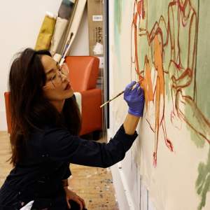 Seonna Hong paints figures onto a canvas tacked to the wall in an art studio. She has long dark hair, light skin tone tone and wears wireframe glasses and a navy jumpsuit.
