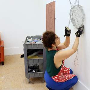 Sarah House, a white woman with light skin tone and dark wavy brown hair pulled up into a bun, crouches in front of a wire sculpture on a white wall in a studio.