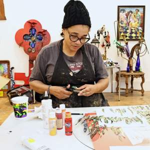 J.Renee works at a table cutting small scraps of painted paper in a studio filled with colorful artwork. She is a Black woman with medium skin tone, hair tucked into a black cap, wearing a paint splattered apron and glasses.