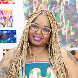 Soraya smiles at the camera, wearing cat eye glasses, a gold septum piercing and gold necklaces. She has long blonde-brown thin braids, and medium dark skin.