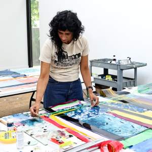 Ivelisse works with colorful fragments of paper and plastic on a work table full of strips. She is a Puerto Rican woman with dark curly hair and medium light skin tone wearing jeans and a graphic tshirt.
