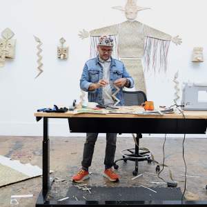 Salvador Jiménez-Flores works with a palm weaving at a work table in front of a white wall hung with various woken masks and figures. A Latino man with medium skin tone, wears denim jacket, jeans, glasses and a baseball hat.