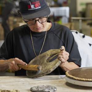 Sahar Khoury uses a wire brush on a textured disc sitting at a studio table. She has dark curly hair under a black Democracy Now baseball hat, wears a black shirt, glasses, gold chain and has a medium-light skin tone and short black painted fingernails.