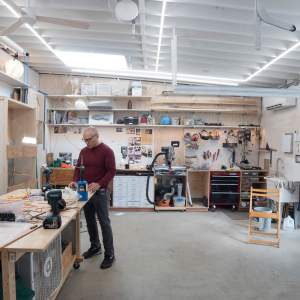 William Lamson, a Latino man with light skin tone, shaved black hair, and black rimmed glasses, stands at a work table in his studio, a garage space with concrete floors, white walls and ceiling, and multiple tools for woodworking and sculpture.