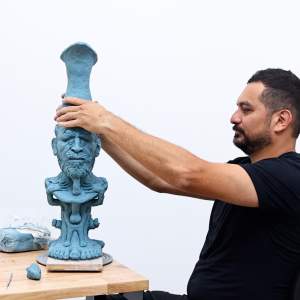 Rodrigo Lara works with his hands placing a crown on a blue male figurative bust in a studio. He is a Mexican man with a dark beard, short dark hair, and wears a black tshirt.