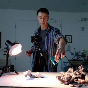 In a darkened studio, Ryan Leitner shines a flashlight with a colored film over an abstract sculpture while photographing it. He is a white man with light skin tone, short dark hair, and a striped button-up.