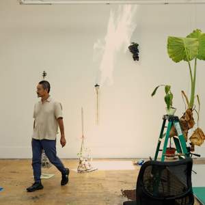 Fred Liang strides through a studio with plants in vials attached to the wall and a banana plant on a ladder. He is a Chinese-American man with short greying hair.