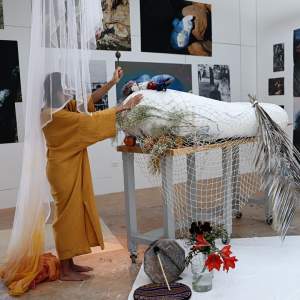 Love Nguyen, a Vietnamese-American woman with long dark hair wearing a mustard shift dress, stands under a white veil with a shaker in her upraised hand.