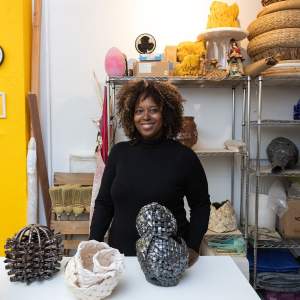 Anina Major, a Black woman with medium-dark skin tone and brown and black curly hair, stands in an artist’s studio behind a table holding her ceramic sculptures. On the metal shelves behind her, we see baskets, other ceramic sculptures, and boxes.