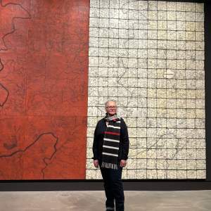 Brenda Mallory stands in front of a large abstract diptych. She is a citizen of Cherokee Nation, with long gray hair pulled back, light skin tone and wearing all black with a wide white and red striped black scarf.