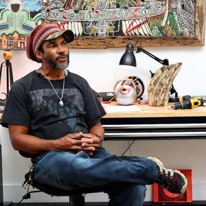 Mankh sits in a chair in a studio with abstract and figurative artwork and sculptures. He is a Black man with medium skin tone, and hair tucked into a knit cap with a greying beard.