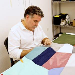 Gabriel Martinez works with a needle and thread at a patchwork quilt on a table. He is a Latino man with light skin tone, greying curly hair and a grey mustache wearing a white button-up shirt.