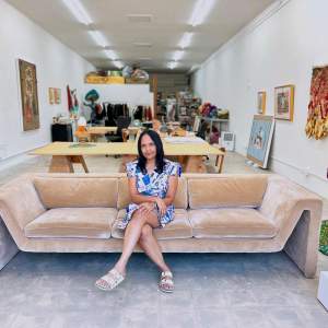Suchitra Mattai sits on a tan couch in a long studio with artwork covering the walls. She is a woman of Indo-Caribbean descent with medium skin tone and long dark hair, wearing a blue floral dress.