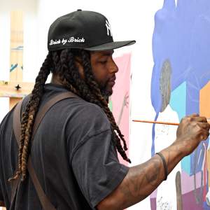 Jay Mckay paints on an unstretched canvas. He is a Black man with long braids and dark beard, wearing a black hat and shirt.
