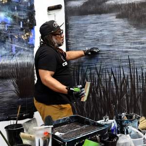 Michi Meko works on a large canvas tacked to the wall, of an abstracted watery landscape. He is a Black man with medium dark skin tone and long dreadlocks, and wears a baseball hat, black t shirt and apron.