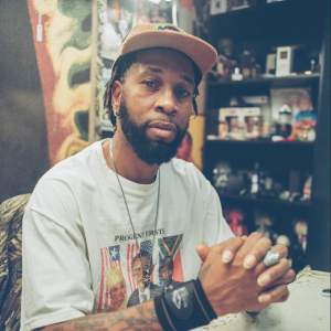 Demond Melancon is seated at a table in an interior space, facing the camera with hands clasped in front of him, resting on the table. He is a Black man with medium dark skin tone, light brown eyes, and a dark short beard and short dreadlocks. He wears a tan corduroy baseball hat, a silver chain with a medallion and a t-shirt with a faded graphic reading “Progeny Firsts” with photos of Sirleaf, Obama, and Mandela.