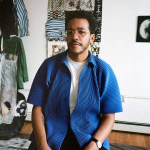 Troy Montes Michie sits in a studio with collage and textiles on the wall behind him. He is a Black and Latino man with short dark hair and a mustache, wearing wireframe glasses and a textured blue shirt.