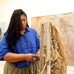 Jessica works on braiding rough twine on a sculpture. She is a Black artist with long curly black hair, and wears a blue button up and olive pants. Behind her is a large abstract painting and a self portrait.