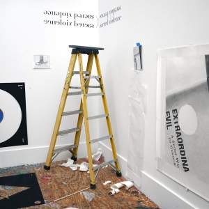 In the corner of a white walled studio with a plywood floor, a 6 foot yellow ladder stands at center, surrounded with various taped prints and papers hanging from the wall. Above the ladder in the corner are the words “sacred violence” mirrored above and to the side , wrapping around the corner. To the left is a black square with a large circle cut in center, and a blue circle within that like a bullseye, with a wooden dowel leaning against the center. To the right is a large print of a close up of a fragment of a page with cut-off text reading “extraordina evil”.