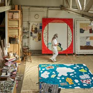In a wide view of an artist's studio with white walls, trestle beam ceiling, and concrete floors covered in dropcloths, artist Rebecca Morris walks in front of a large painting, wearing a white jumpsuit and respirator, and wiping her hands with a cloth. Her paintings rest against the walls and floor and are stored on shelving.