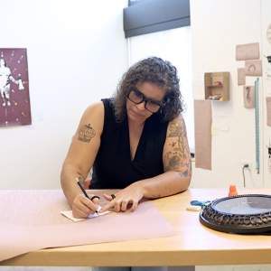 Lilliam Nieves, a Puerto Rican woman with medium light skin and black and grey curly hair, leans over a desk writing on brown paper. She wears chunky glasses, a black top and has several tattoos.