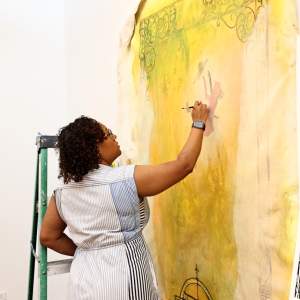 Nadrea paints on a large yellow canvas tacked to the wall. She is a Black woman with shoulder length black braids, and wears glasses and a striped dress.