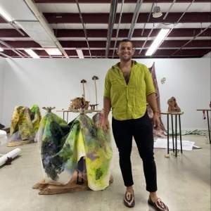 The artist Javier Orfon, a Puerto Rican person with medium skin tone, dark short hair, and face stubble, stands in a room with sculptural artworks behind him. He smiles at the camera, wearing a bright green shirt, left open at the top, black pants, and snakeskin loafers.