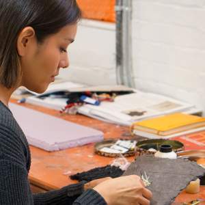 Mikayla Patton, an Oglala Lakota and Isleta Pueblo woman with medium light skin tone and chin-length straight dark hair, sits at an orange worktable, facing partially toward a white brick wall. She works with porcupine quills and handmade paper to create a sculpture. Various books, tools, and other materials sit on the desk.