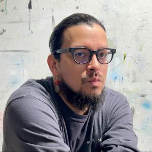 A Ho Chunk Native American with medium-light skin and a beard faces the camera, wearing black framed glasses and a beaded earring. The backdrop is a white wall in his art studio, with drips and splashes of paint visible on the wall.