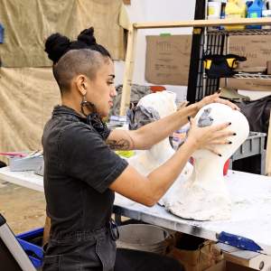Maya Pen sits at a work table shaping clay onto a bust sculpture. She is a latina woman with the sides of her head shaved, and dark hair pulled into buns on top of her head, wearing large earrings and a work jumpsuit.