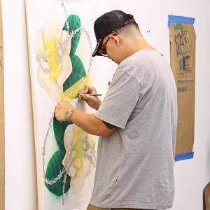 Angel Perdomo, a Latino man wearing glasses and a baseball hat, works on a painting in a white-walled studio.