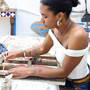 Ute Petit draws on a mixed media piece with a wooden frame, surrounded by art supplies and work in her studio. She is a Black woman with a medium skin tone. wearing an off the shoulder white shirt, a cross necklace and hoop earrings, with her dark hair pulled back in a bun.