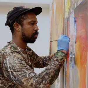 Pat Phillips, a Black man with medium-dark skin, a dark beard, wearing a baseball hat and a camoflage patterned shirt, paints on a canvas on a wall.