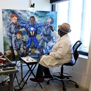 Antoine Prince works on a large oil painting of a Black soldier in Captain American uniform holding up bodies of German soldiers. Antoine is a Black man, wearing a white painters coar, and a brimmed hat.