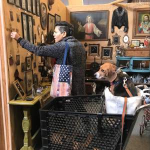 In a stall of an antiques mall, Sara reaches for a crucifix on a temporary wall densely hung with pictures and icons. Sara has light-toned skin and short black hair, and she wears a heathered gray sweater. A beagle sits in a bag in the basket of a shopping cart between us and the artist.