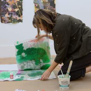 Artist Naomi Kawanishi Reis kneels over a pile of paper with green watercolor washes on the floor of a studio. She is an artist of Asian descent, and has brown shoulder-length hair with blonde streaks and bangs, and wears a green putton up shirt, dark pants and pink socks.