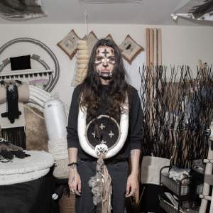 Eric-Paul Riege stands in a room with artwork and materials. He is a Diné/Navajo man with long dark hair, and wears black and white paint with a bead and wire jewelry across his nose. Around his neck is a thick white coil with a large black medallion with 4 crosses.