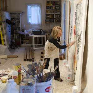 In an art studio, a Latina woman with light skin tone and shoulder-length blond hair leans towards a canvas painting leaning on the wall, her hand moving across the surface with a paintbrush. In the foreground are cans and buckets filled with paint brushes, and a table with tubes of paint.