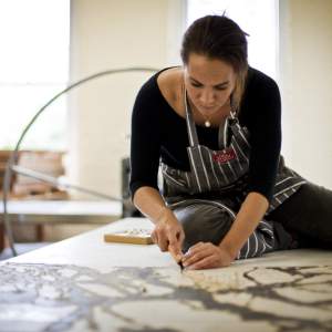 Abigail Romanchak kneels on a table, working on a large abstract drawing with dark lines and shapes, in a room with art supplies and an etching press in the background. She is a Hawaiian multi-racial woman with medium skin tone and dark long hair, and wears black clothing under a striped apron.