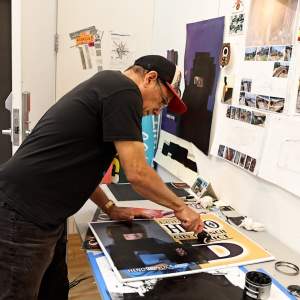 Chemi Rosado-Seijo, a Puerto-Rican man with medium skintone, wearing a baseball hat and black tshirt and jeans, uses a ink brayer to roll black onto a political yard sign.