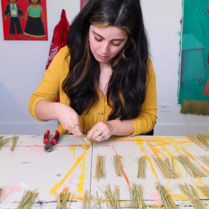 Karla works with bundles of straw in her studio. She has light skin, long dark hair, and a sunflower yellow shirt.
