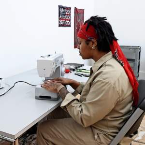 Sean work at a sewing machine. She is a black artist with short black hair, pulled back with a red sash. She has tattooed forearms, and wears a khaki worksuit.