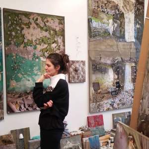 In an artist’s studio, a white woman with brown hair, a white turtleneck, and black sweater and pants touches her face with her hand while looking toward the left frame of the photo. On the wall are paintings of distressed interior spaces. Small paintings and art making materials surround the artist on the floor.