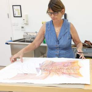 Katy Schimert, a white woman with bobbed blond hair, adjusts an abstract draing on a table. She wears glasses and a denim sleeveless dress.