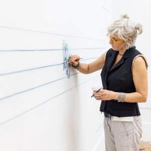 Analia Segal paints with blue watercolor a looping abstracted word form directly on a white studio wall. She is an Argentine-American artist with grey hair pulled up into two small ponytails, light skin tone, and wears a black vest, lined pants, and silver chain bracelets.