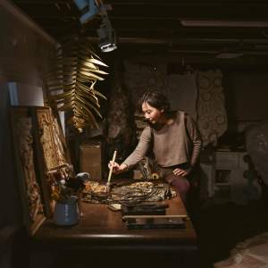 Sammy Seung-min Lee is of Korean descent and has straight black hair and medium-toned skin. Lit only by a spotlight in an otherwise dim studio, she perches one hip on the edge of a table as she paints using a calligraphy brush with the other.