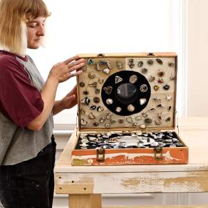 Sadie Sheldon opens a briefcase filled with arranged silver-encased objects. She is a white person with light skin tone with short blond hair.